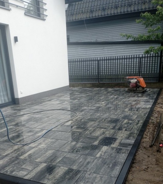 Ułożona, mokra kostka brukowa w odcieniach szarości z czarną obwódką, widoczny wąż i zagęszczarka w tle przy ogrodzeniu.