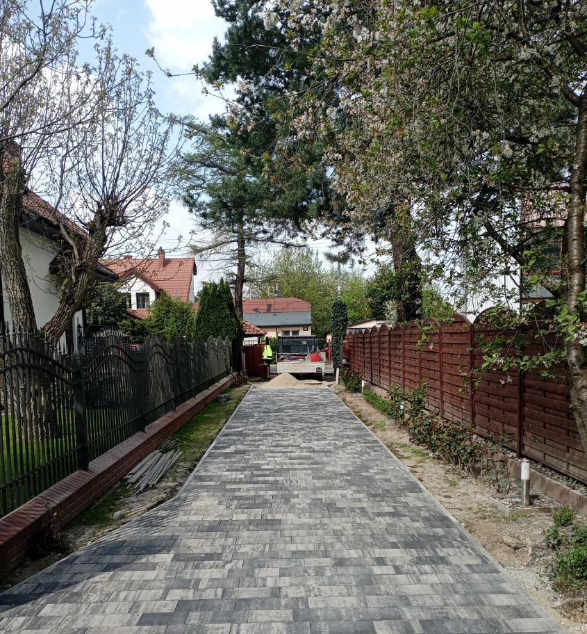 Długa, wybrukowana droga dojazdowa wykonana z szaro-czarnej kostki, otoczona ogrodzeniami z drewna i metalu, z widoczną ciężarówką w oddali i pracownikiem w kamizelce odblaskowej.