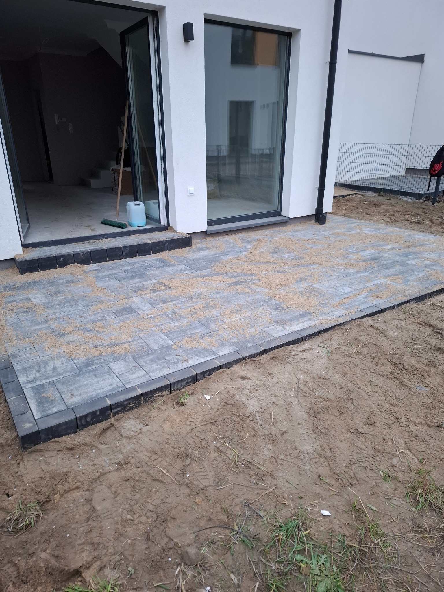 Nowo ułożony taras z szarej kostki brukowej, z czarnym obramowaniem, posypany piaskiem, przylegający do białego budynku z dużymi szklanymi drzwiami tarasowymi.