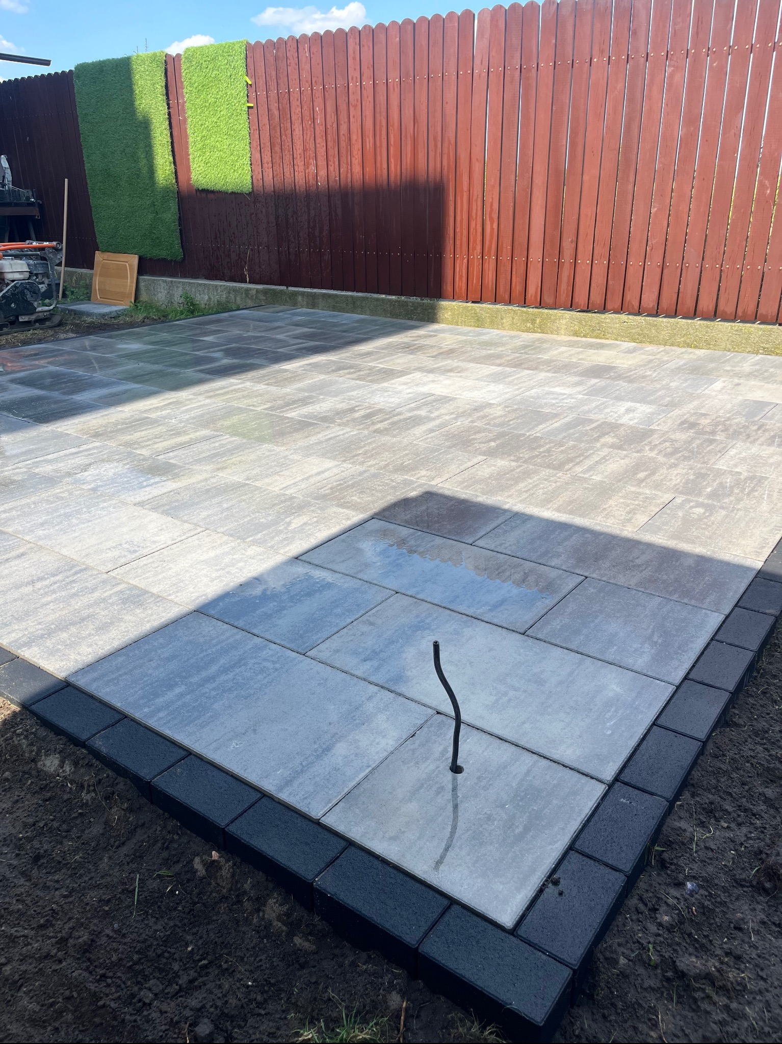 Ułożona szara kostka brukowa z czarnym obrzeżem przy brązowym płocie z dekoracyjną sztuczną trawą w tle. W kostce zamontowany element instalacji.