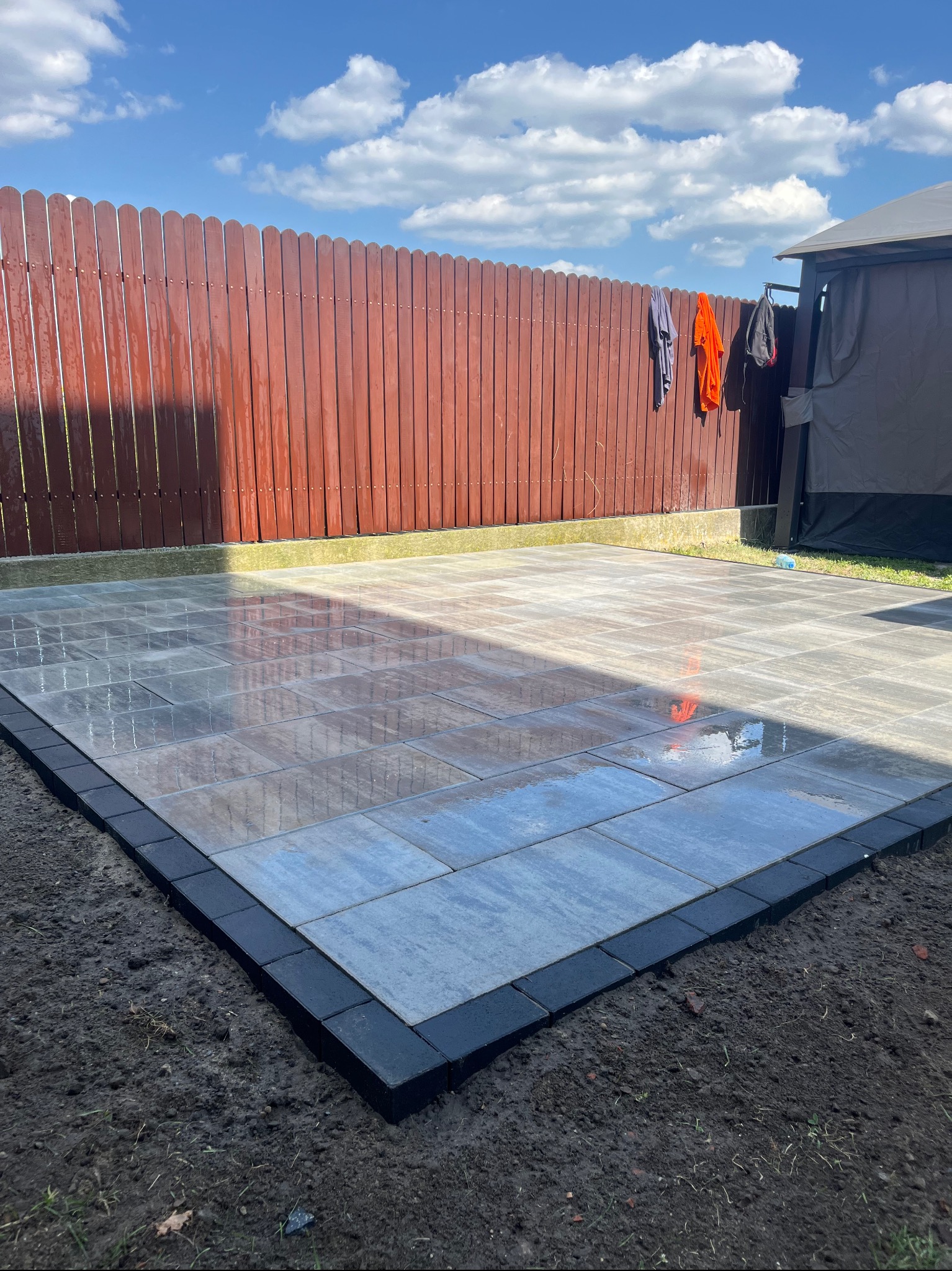 Ułożona, wilgotna kostka brukowa w ogrodzie, otoczona czarnym obramowaniem. W tle brązowe ogrodzenie i fragment altany. Na płocie wiszą ubrania.