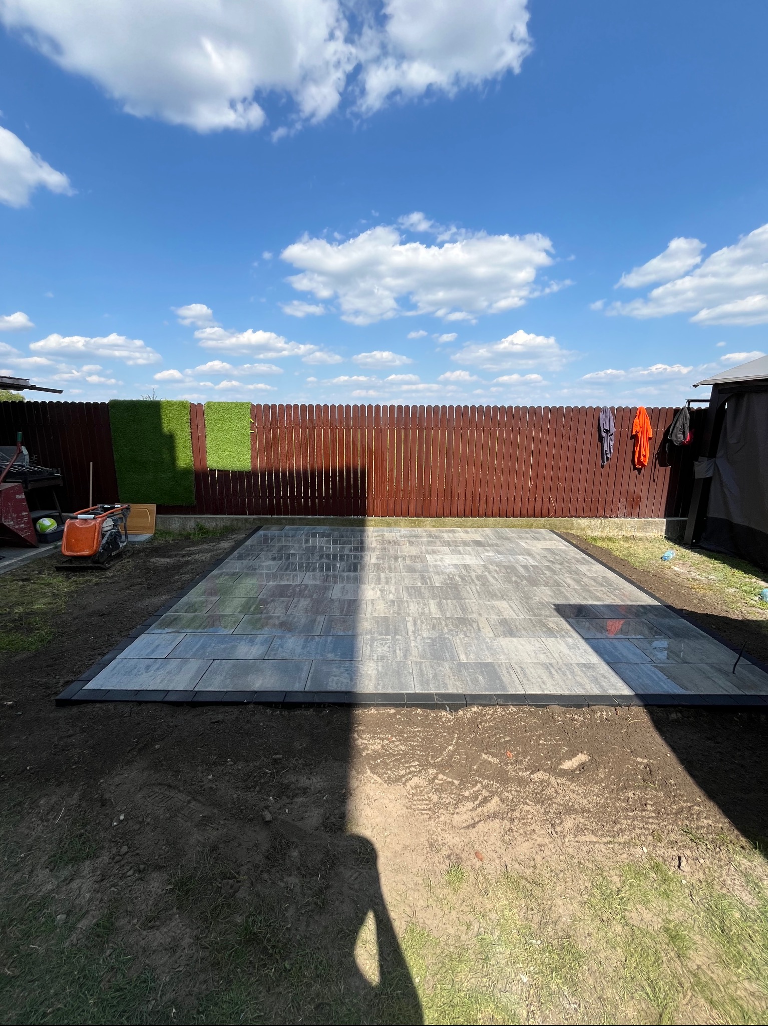 Świeżo ułożona, szara kostka brukowa z czarnym obramowaniem na przygotowanym podłożu. W tle brązowe ogrodzenie i błękitne niebo z chmurami.