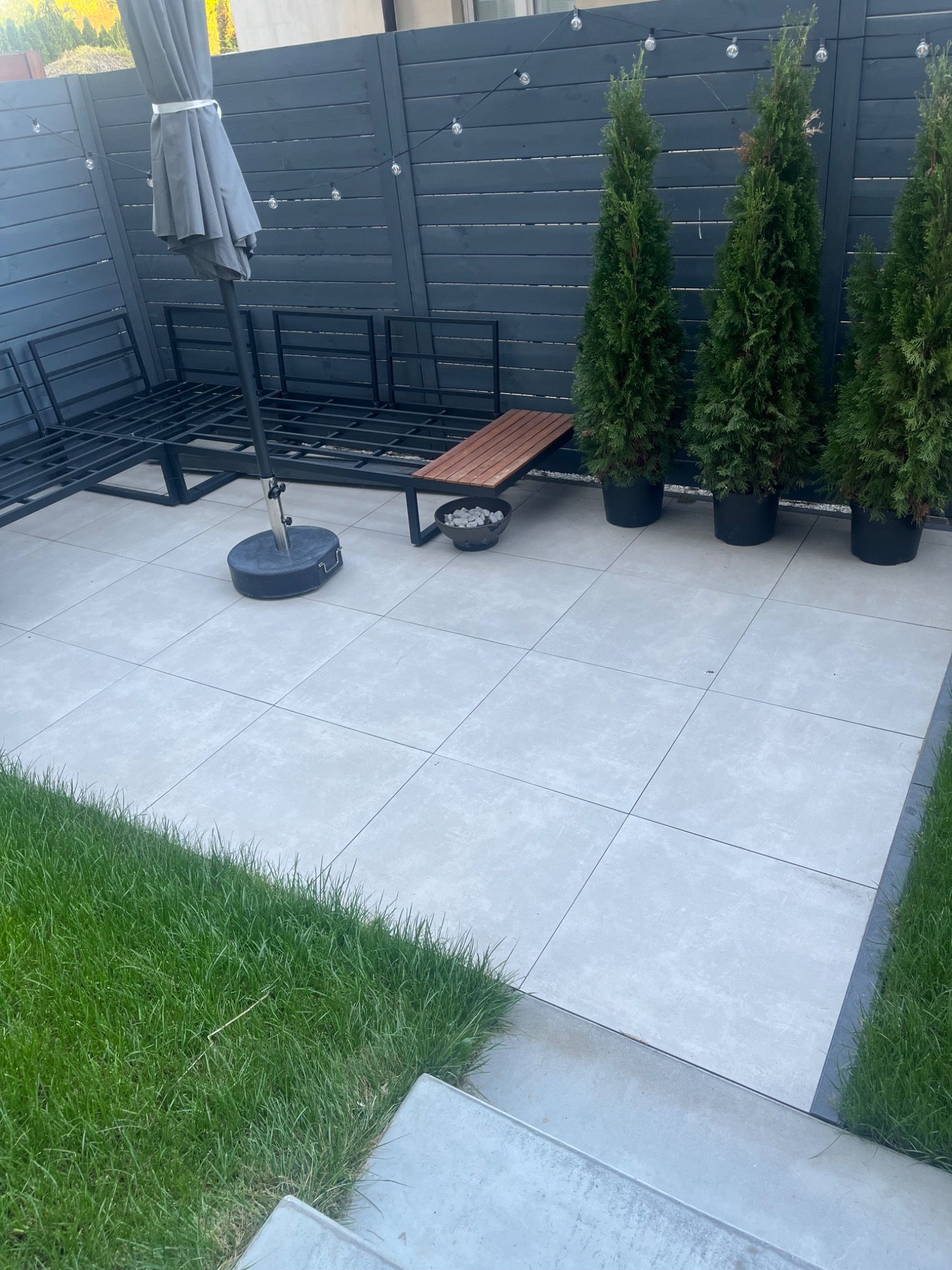 Jasnoszare płytki tarasowe ułożone w kwadraty, z elementami wyposażenia: parasol, sofa ogrodowa i tuje. Tło stanowi ciemnoszary płot z lampkami.