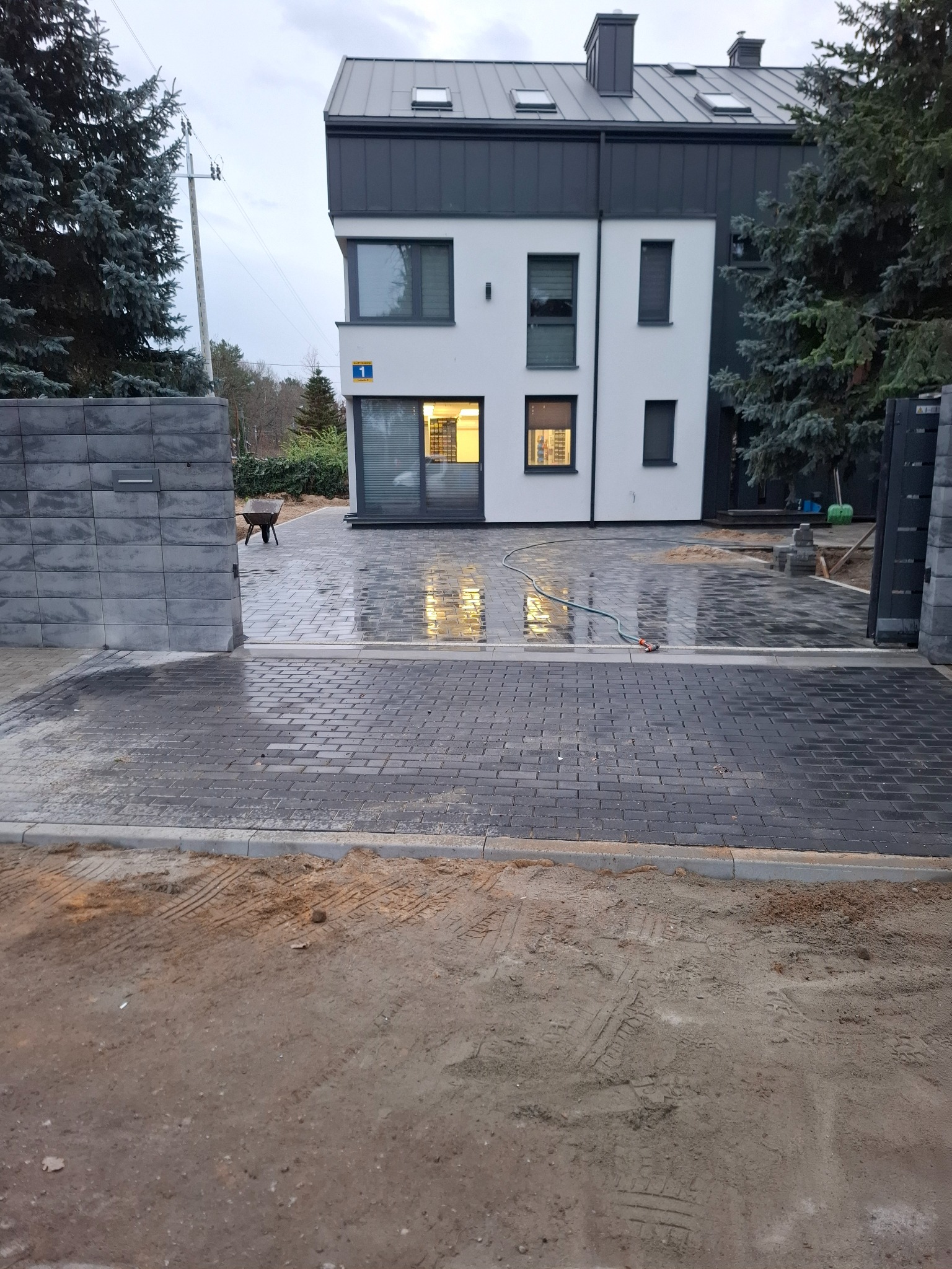 Nowoczesny podjazd z szarej kostki brukowej prowadzący do domu z numerem 1, widoczny fragment ogrodu i brama wjazdowa. Świeżo ułożona kostka, mokra od wody.