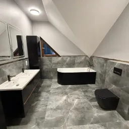 Nowoczesna łazienka na poddaszu z szarymi marmurowymi płytkami, czarną wanną wolnostojącą, umywalką z dwoma kranami i wiszącą toaletą.