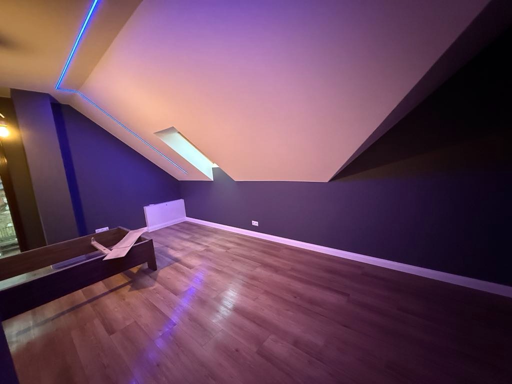 Poddasze po remoncie: ściany pomalowane na ciemny fiolet, drewnopodobne panele na podłodze, białe listwy przypodłogowe, okno dachowe, niebieskie oświetlenie LED przy suficie, częściowo widoczny...