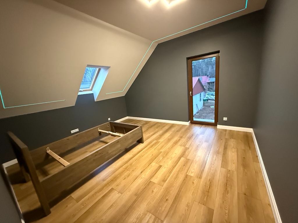 Pokój na poddaszu po remoncie, z szarą ścianą, drewnianą podłogą, oknem dachowym i ramą łóżka bez materaca. Widoczna linia lasera na ścianach i suficie.