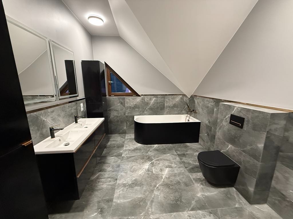 Nowoczesna łazienka na poddaszu z szarymi marmurowymi płytkami, czarną wanną wolnostojącą, umywalką z dwoma kranami i wiszącą toaletą.