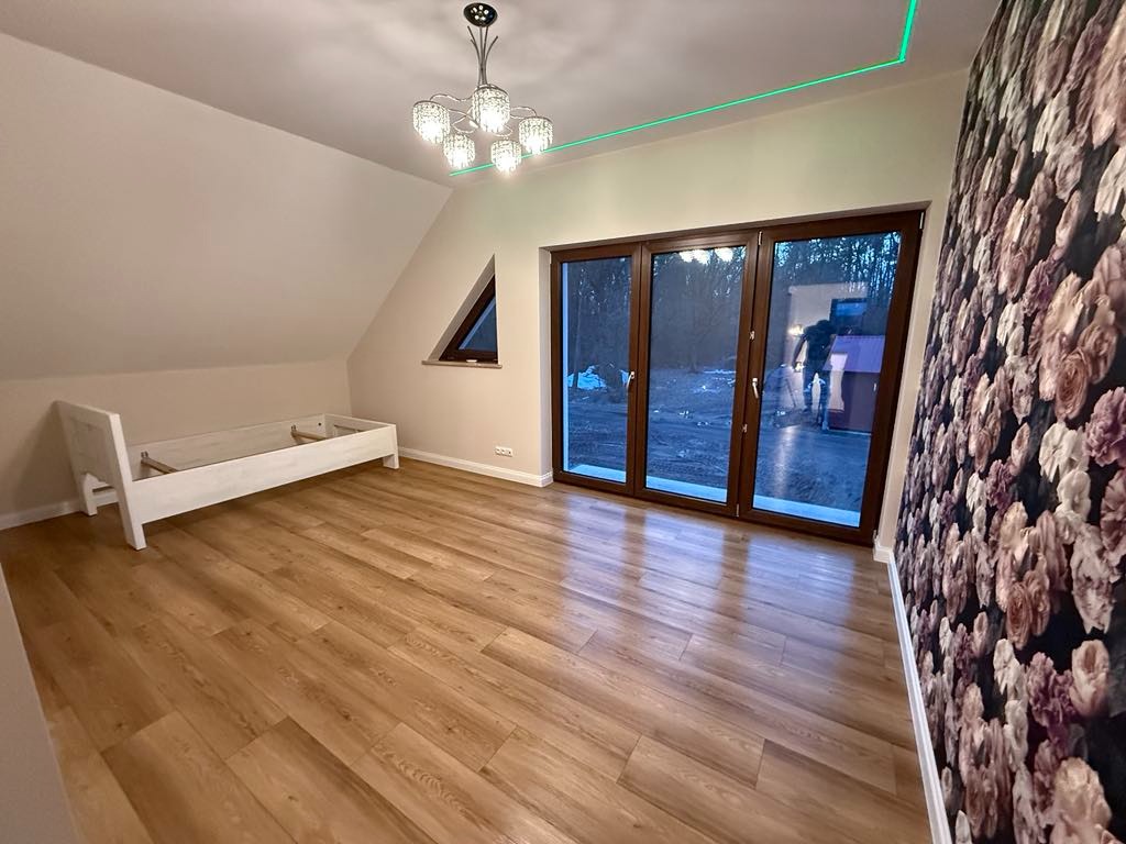 Pokój na poddaszu po remoncie: drewniana podłoga, białe ściany, okno trójkątne, duże okno balkonowe, designerski żyrandol i ściana z tapetą w kwiaty.