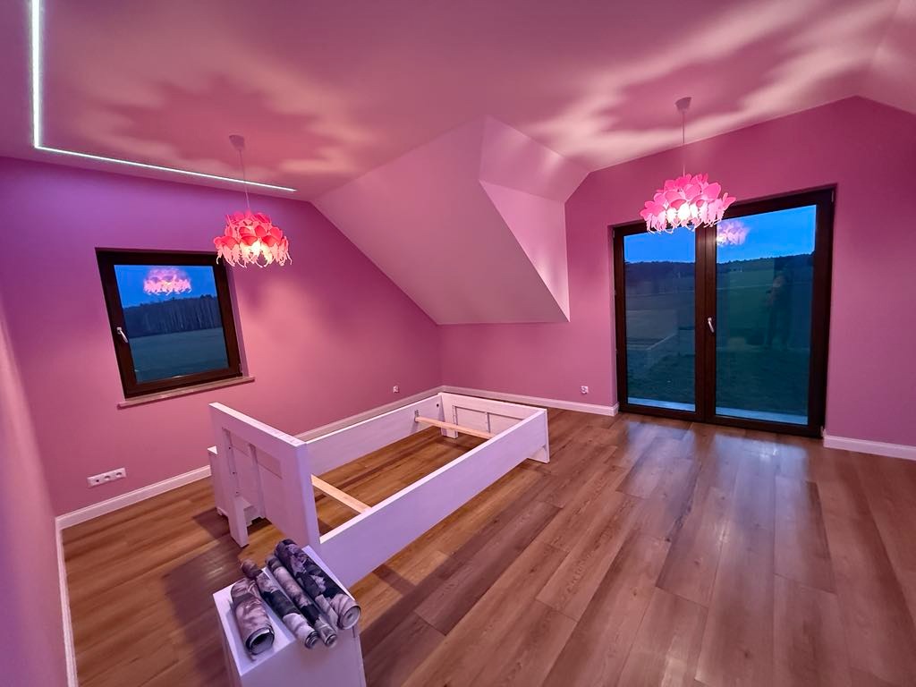 Pokój w odcieniach różu z drewnianą podłogą, białym łóżkiem bez materaca i zwisającymi lampami w kształcie kwiatów. Na podłodze leżą rolki tapety na tle świeżo pomalowanych ścian.