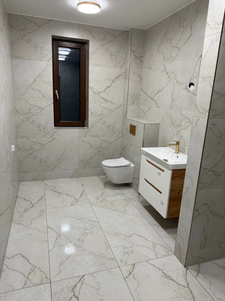 Wykończona łazienka z płytkami imitującymi marmur, podwieszana toaleta, biała szafka z drewnianymi bokami i złotą armaturą, małe okno z ciemną ramą.