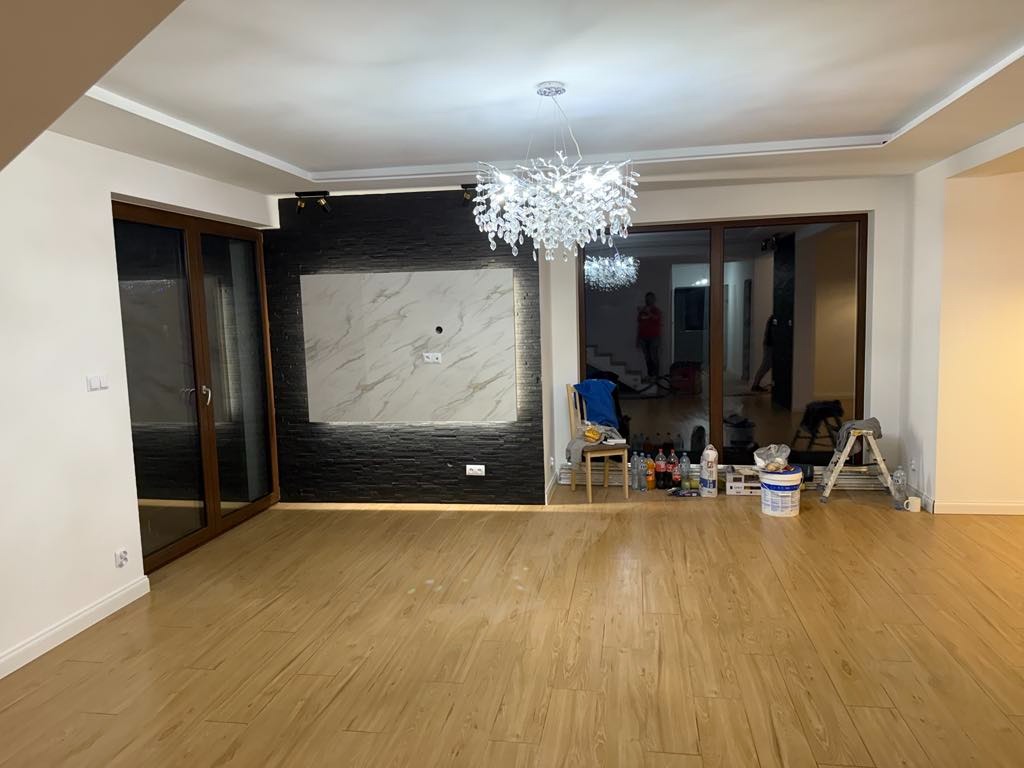 Przestronny salon w trakcie prac wykończeniowych: drewnopodobne panele podłogowe, czarna ściana z dekoracyjnym panelem imitującym marmur, ozdobny żyrandol, widoczne narzędzia i materiały malarskie,...
