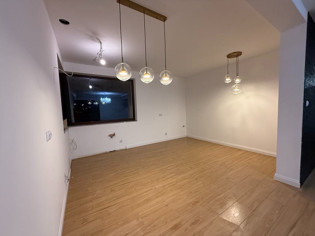 Puste pomieszczenie z drewnopodobną podłogą, świeżo pomalowanymi białymi ścianami, trzema wiszącymi lampami z kulistymi kloszami i widocznymi niedoskonałościami na ścianach w okolicy okna.