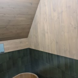 FK DREW - Drewniana wanna w rogu pomieszczenia z panelami ściennymi w kolorze jasnego drewna i ciemnozielonymi płytkami.