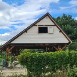 FK DREW - Drewniana konstrukcja altany z białymi panelami i oknem z okiennicą, widoczny napis 'A.D. 2022' na szczycie, częściowo zasłonięta przez zielony żywopłot, na tle błękitnego nieba z chmurami.