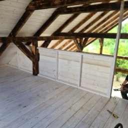 FK DREW - Wnętrze altany w trakcie budowy, widoczne drewniane belki stropowe, białe deski podłogowe i fragment barierki, na podłodze leży nóż monterski.