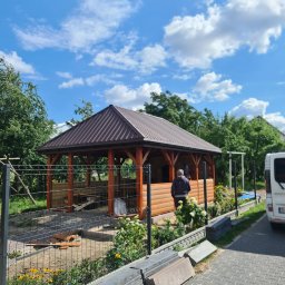 FK DREW - Nowa drewniana altana z brązowym dachem, częściowo osłonięta panelami, widoczna zza metalowego ogrodzenia, z elementami ogrodu i zaparkowanym samochodem w tle.