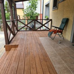 FK DREW - Zadaszony taras z brązowych desek kompozytowych, z drewnianą balustradą, krzesłem ogrodowym i fragmentem elewacji budynku w tle.