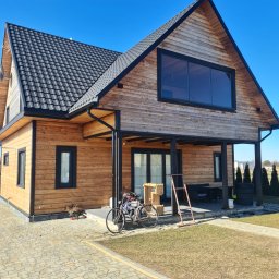 FK DREW - Nowoczesny, drewniany dom z czarną stolarką okienną i dachem z dachówki, widziany z perspektywy chodnika z kostki brukowej w słoneczny dzień. Rower oparty o ścianę, drabina i drewniane skrzynki...