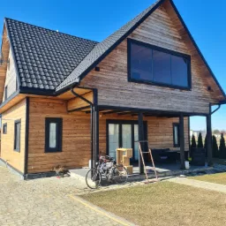 Nowoczesny, drewniany dom z czarną stolarką okienną i dachem z dachówki, widziany z perspektywy chodnika z kostki brukowej w słoneczny dzień. Rower oparty o ścianę, drabina i drewniane skrzynki...