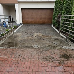 FK DREW - Mokry podjazd z kostki brukowej przed domem z brązową bramą garażową, widoczne ślady po myciu ciśnieniowym, otwarte metalowe ogrodzenie, w tle roślinność.