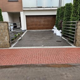 FK DREW - Nowo ułożona kostka brukowa w odcieniach szarości i czerwieni przed domem z brązową bramą garażową, widoczne worki z pozostałościami po budowie i metalowa brama wjazdowa.