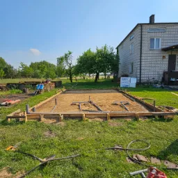 Przygotowany teren pod budowę: drewniany szalunek, piasek, rury kanalizacyjne, narzędzia i worki cementu na trawie, dom w tle.