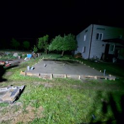 FK DREW - Wylana płyta fundamentowa pod budynek mieszkalny, otoczona drewnianym szalunkiem, widoczna w nocnym oświetleniu, z elementami instalacji wystającymi z betonu.