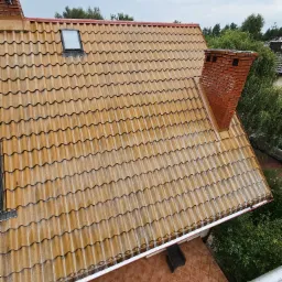 Ceglasty dach z kominem i oknem dachowym po deszczu, widok z góry na mokrą dachówkę, rynny i fragment tarasu z krzesłem.