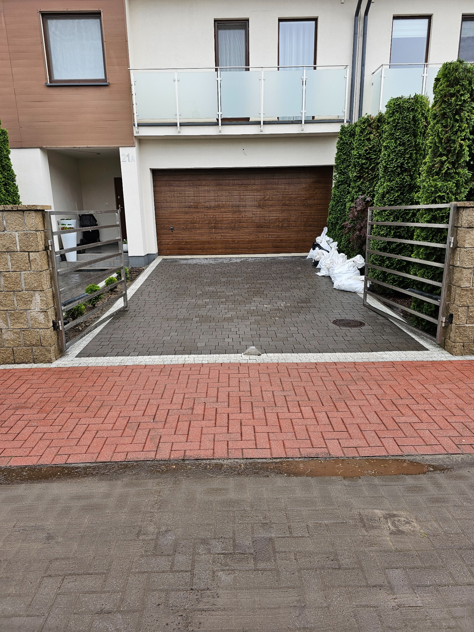 Nowo ułożona kostka brukowa w odcieniach szarości i czerwieni przed domem z brązową bramą garażową, widoczne worki z pozostałościami po budowie i metalowa brama wjazdowa.