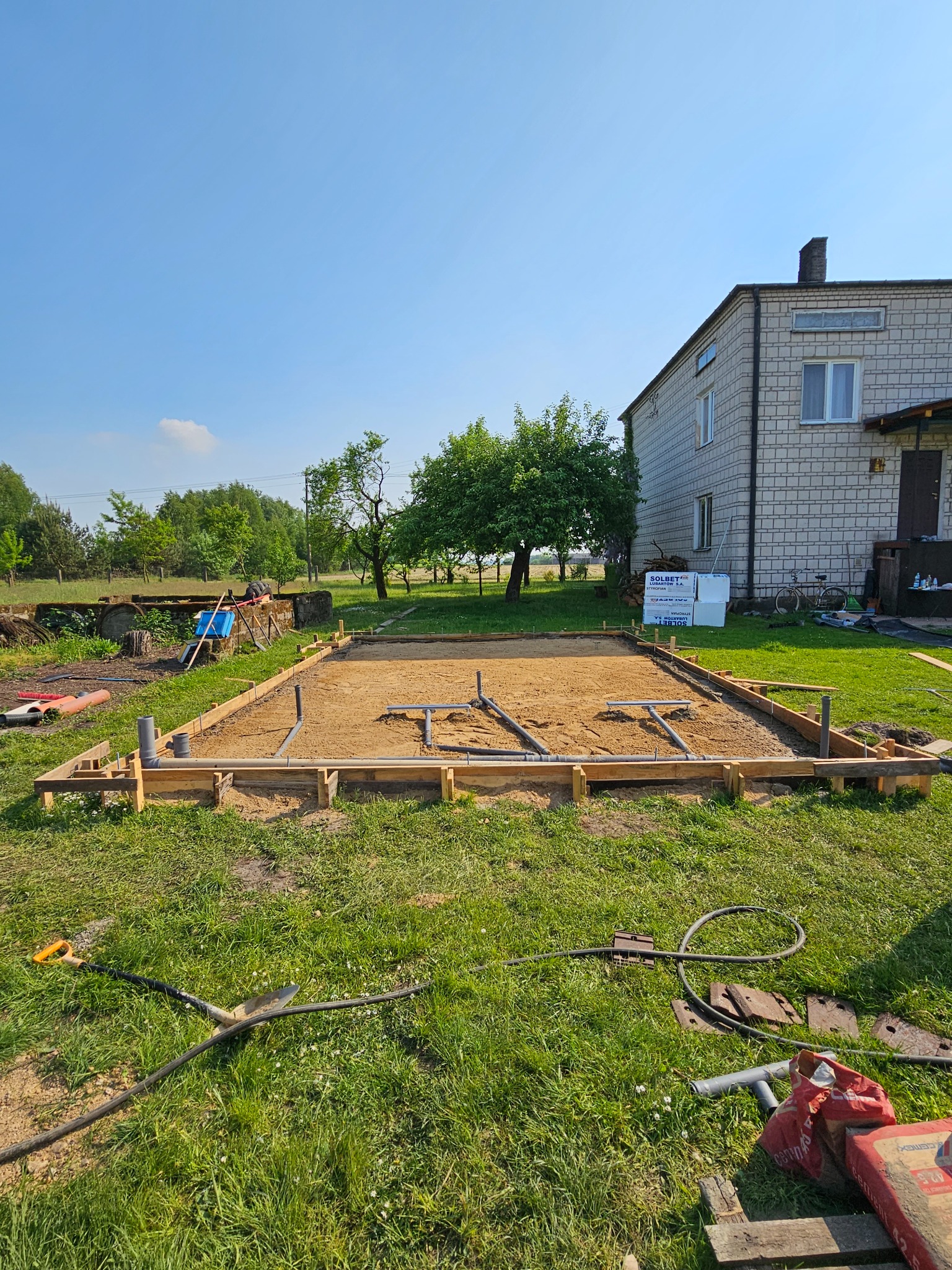Przygotowany teren pod budowę: drewniany szalunek, piasek, rury kanalizacyjne, narzędzia i worki cementu na trawie, dom w tle.