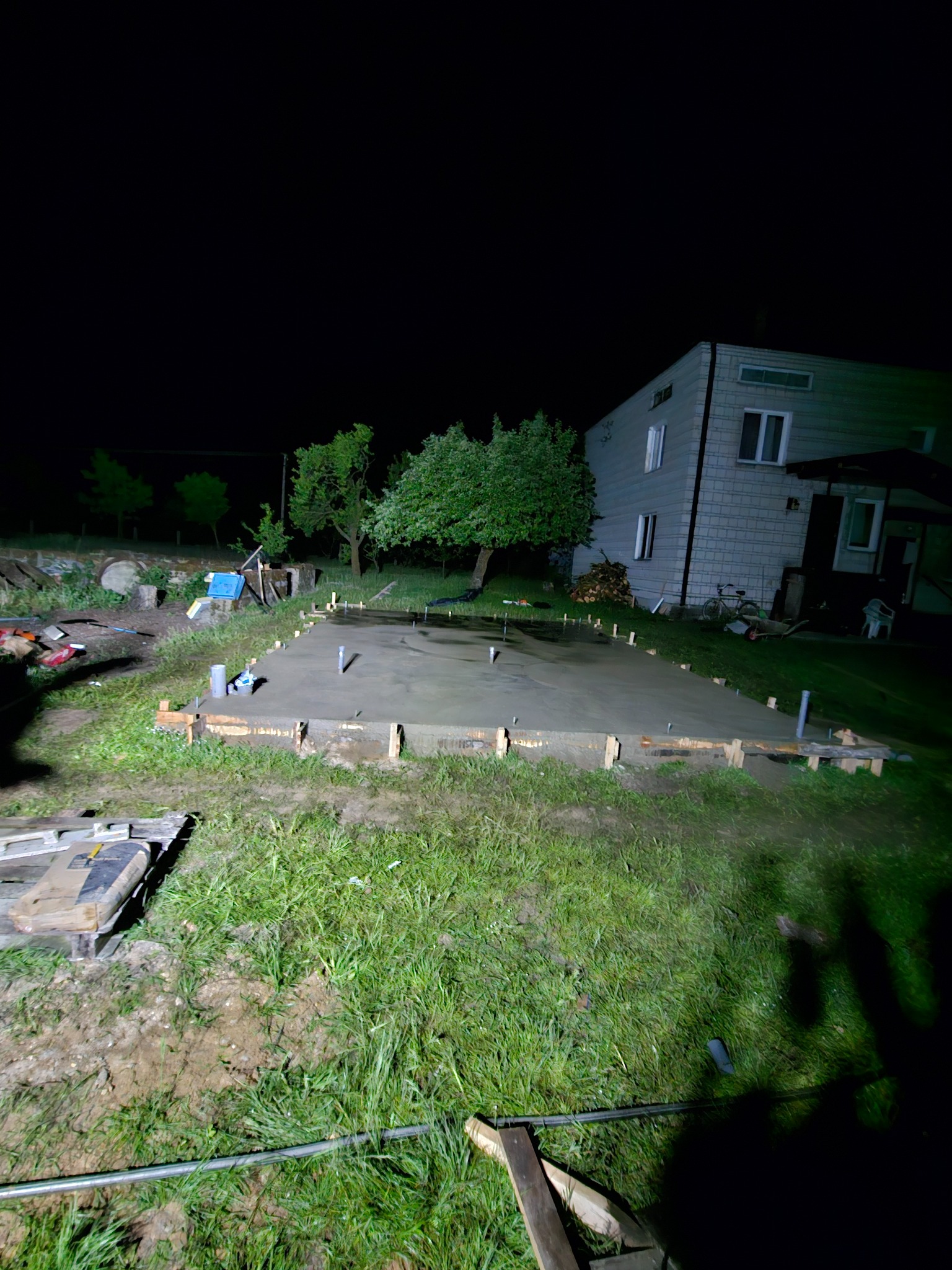 Wylana płyta fundamentowa pod budynek mieszkalny, otoczona drewnianym szalunkiem, widoczna w nocnym oświetleniu, z elementami instalacji wystającymi z betonu.