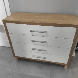 Komoda z czterema białymi szufladami i blatem w kolorze jasnego drewna, z metalowymi uchwytami, ustawiona na szarej podłodze z płytek.