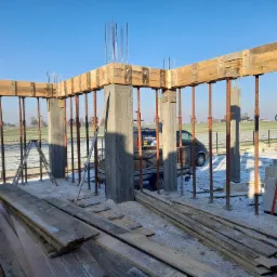 Wznoszenie konstrukcji żelbetowej: szalunek drewniany wsparty na metalowych stemplach, widoczne zbrojenie pionowe, w tle ośnieżone pole i błękitne niebo.