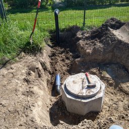 Usługi Hydrauliczne HydroTom - Betonowy zbiornik z pokrywą, młotek i fragment rury w wykopie, Kamień Pomorski. W tle trawa i ogrodzenie.