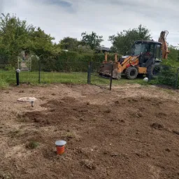 Teren po częściowej niwelacji z widocznymi elementami instalacji sanitarnej i koparko-ładowarką Case w tle, ogrodzenie posesji i drzewa.