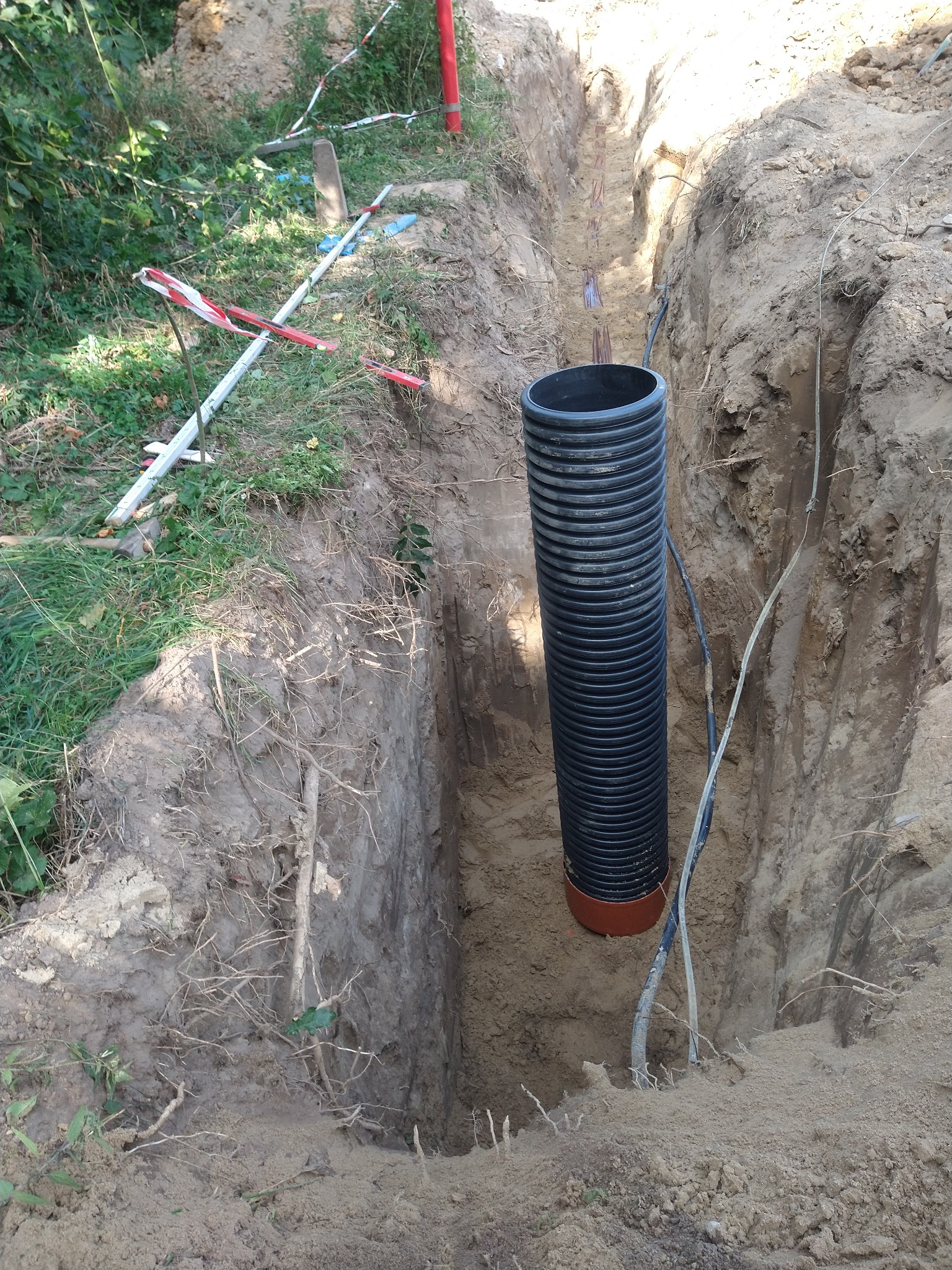 Głęboki wykop w ziemi z widoczną czarną, karbowaną rurą kanalizacyjną z pomarańczowym łącznikiem na dole oraz kablami biegnącymi wzdłuż ściany wykopu. Na powierzchni ziemi widoczne elementy taśmy...