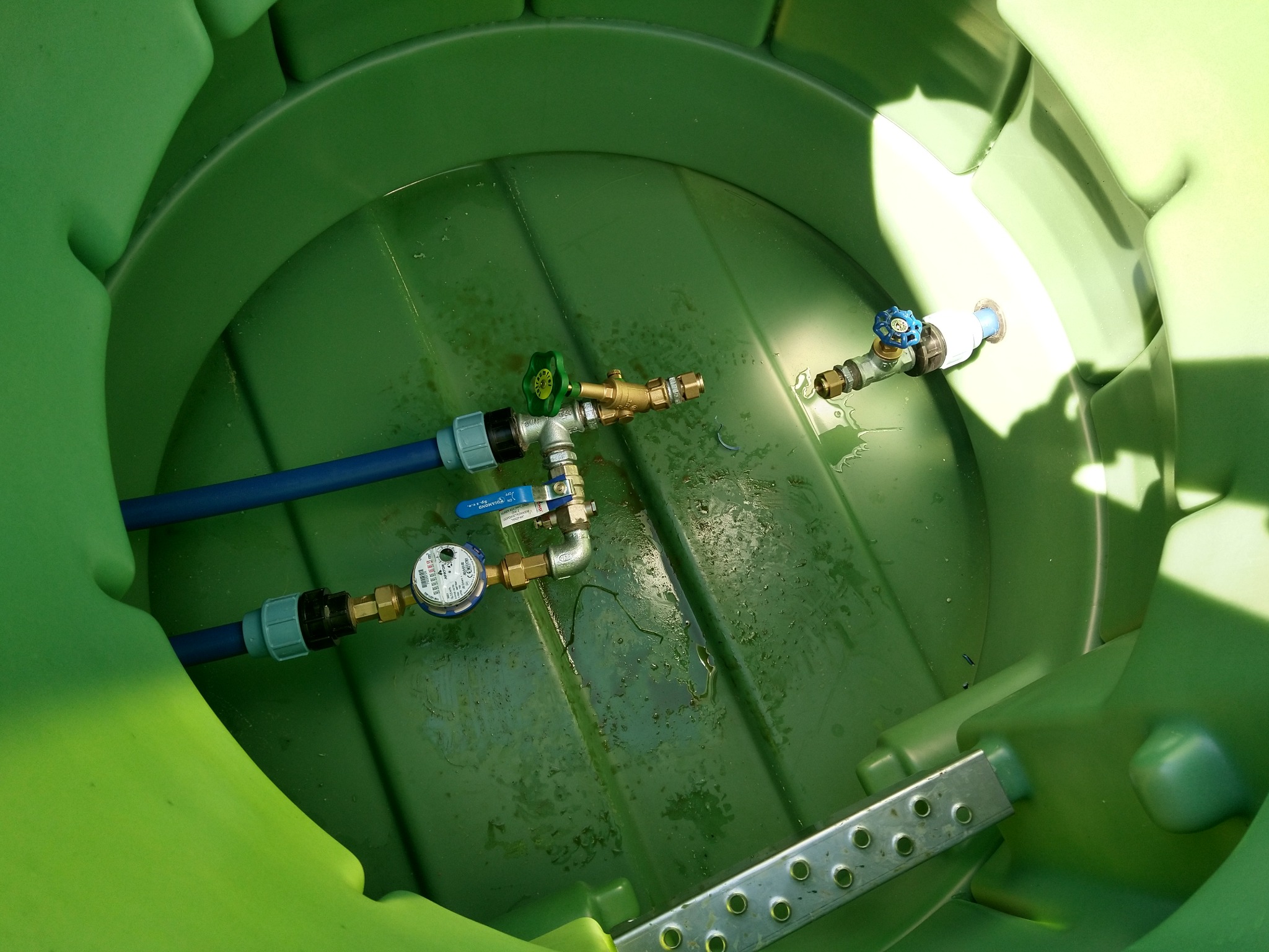 Wnętrze zielonego zbiornika z zainstalowanymi zaworami, rurami i licznikiem wody, widoczne ślady wilgoci na dnie.