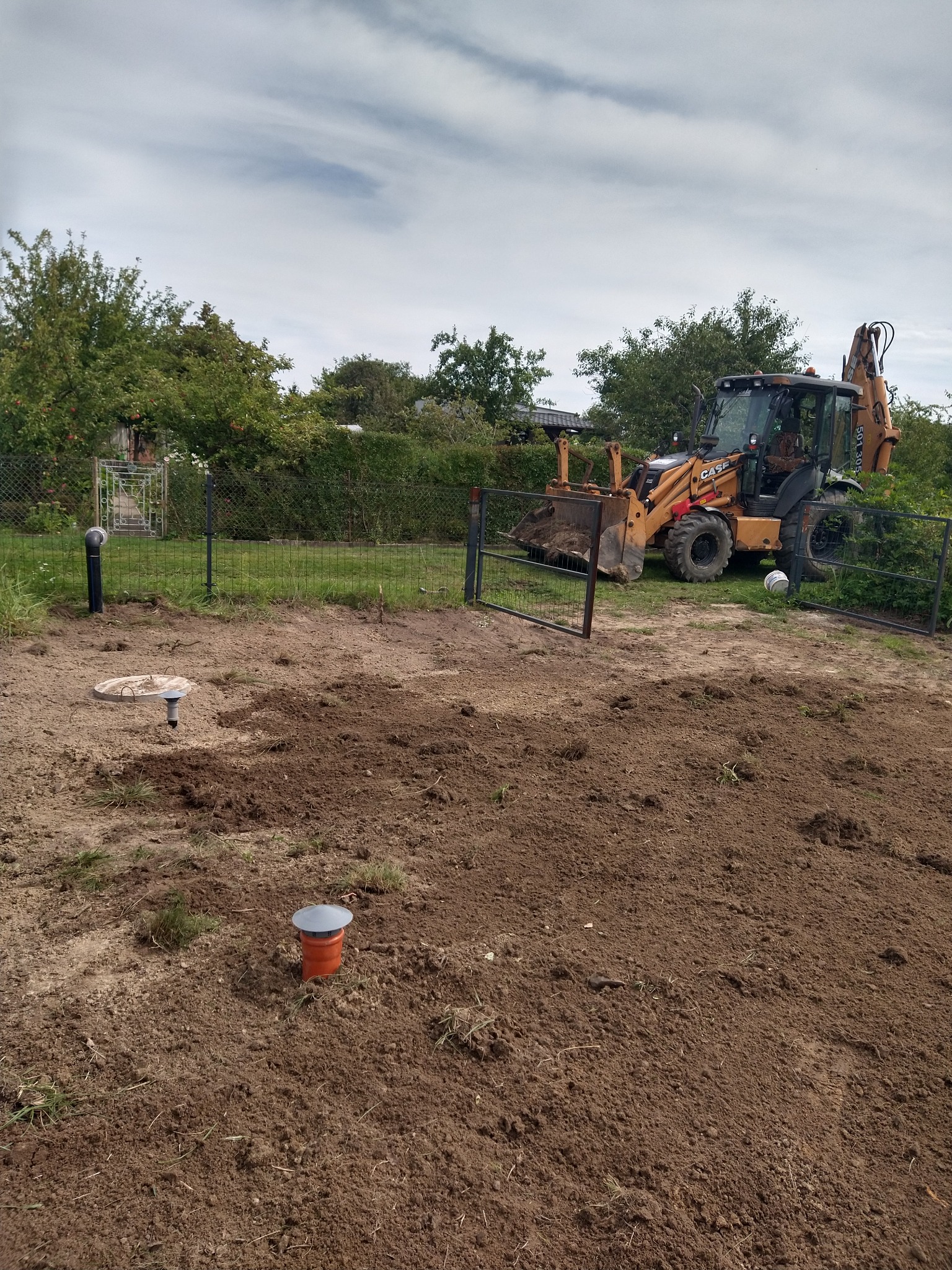 Teren po częściowej niwelacji z widocznymi elementami instalacji sanitarnej i koparko-ładowarką Case w tle, ogrodzenie posesji i drzewa.