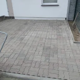 Ułożona kostka brukowa w kształcie litery I, z widocznym włazem kanalizacyjnym i fragmentem ściany budynku z oknem oraz zabezpieczonym kartonem otworem wentylacyjnym.