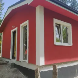 Świeżo pomalowany na czerwono budynek z białymi obramowaniami okien i drzwi, widoczne fundamenty z izolacją, surowe otoczenie placu budowy.