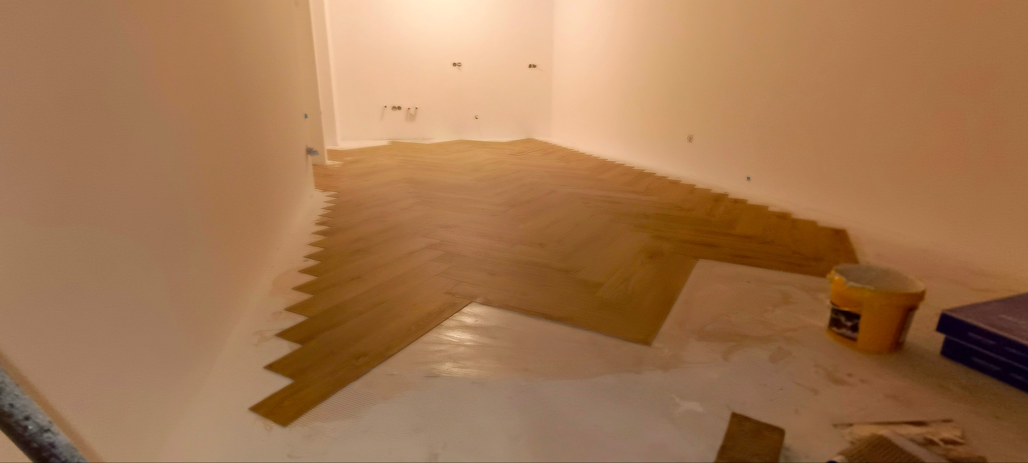 Układanie parkietu w jodełkę w pustym pomieszczeniu z widocznym klejem i żółtym wiadrem po prawej stronie.