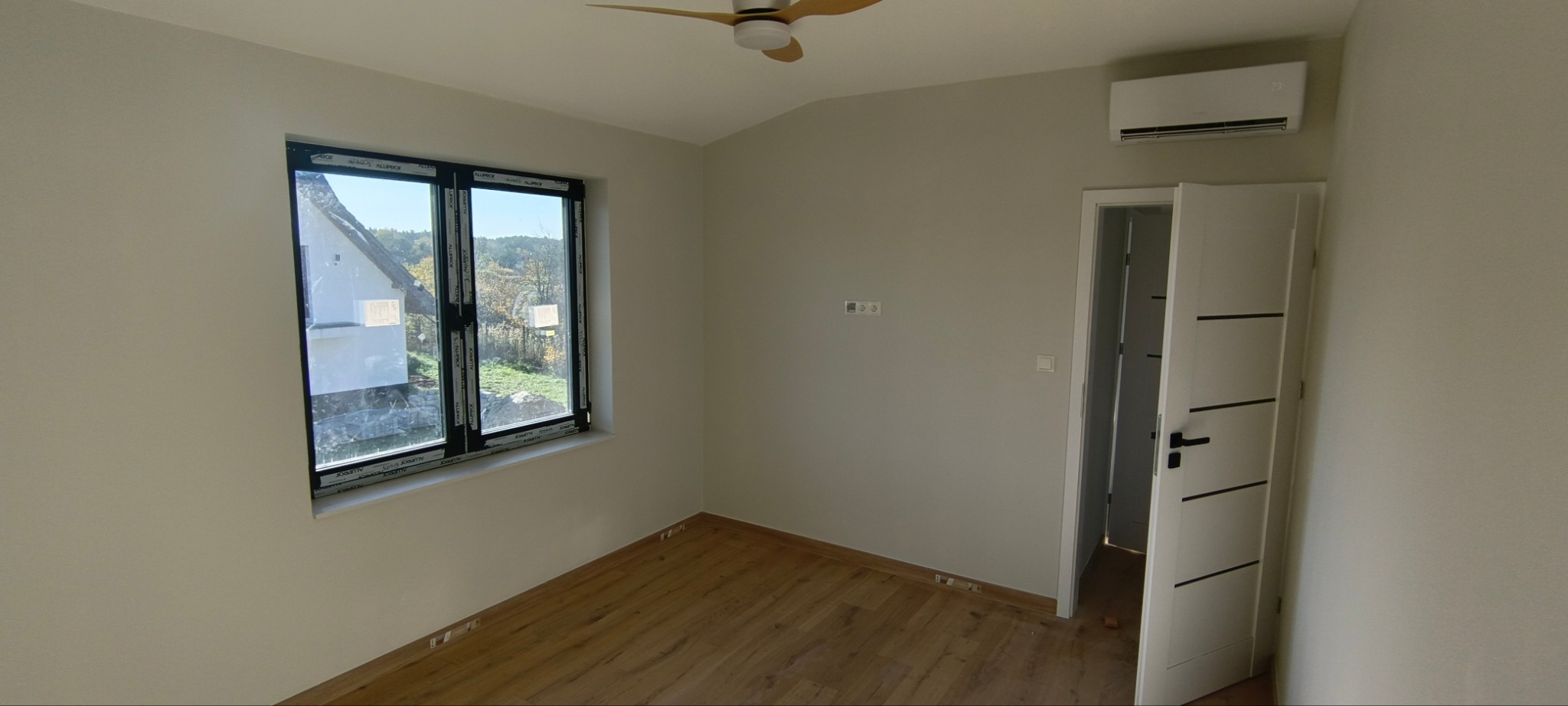 Jasne wnętrze pokoju po remoncie: parkiet, białe ściany, białe drzwi z czarnymi paskami, okno z widokiem na dom i drzewa, klimatyzator na ścianie, wentylator sufitowy.