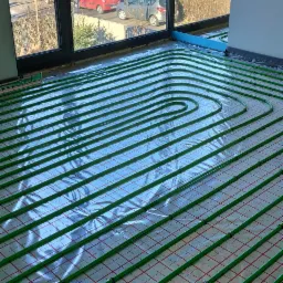 Zielone rury ogrzewania podłogowego ułożone na folii izolacyjnej z czerwoną siatką, widok z góry na tle okna z widokiem na domy i drzewa.