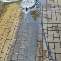 Czyszczenie kostki brukowej za pomocą myjki ciśnieniowej Kärcher, widoczny efekt czyszczenia: po lewej stronie kostka jest czysta i mokra, po prawej brudna i sucha. Na pierwszym planie niebieski...