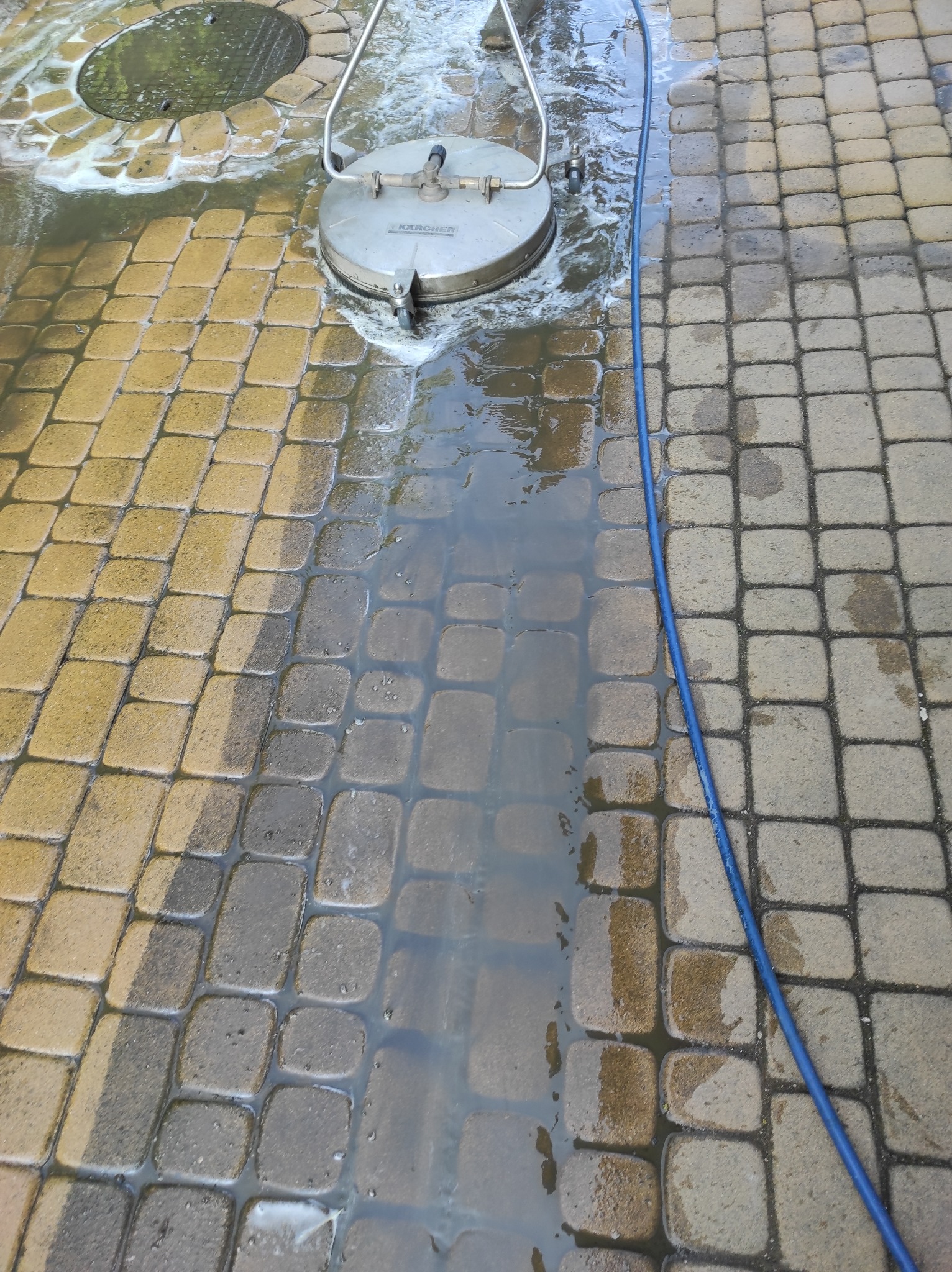 Czyszczenie kostki brukowej za pomocą myjki ciśnieniowej Kärcher, widoczny efekt czyszczenia: po lewej stronie kostka jest czysta i mokra, po prawej brudna i sucha. Na pierwszym planie niebieski...