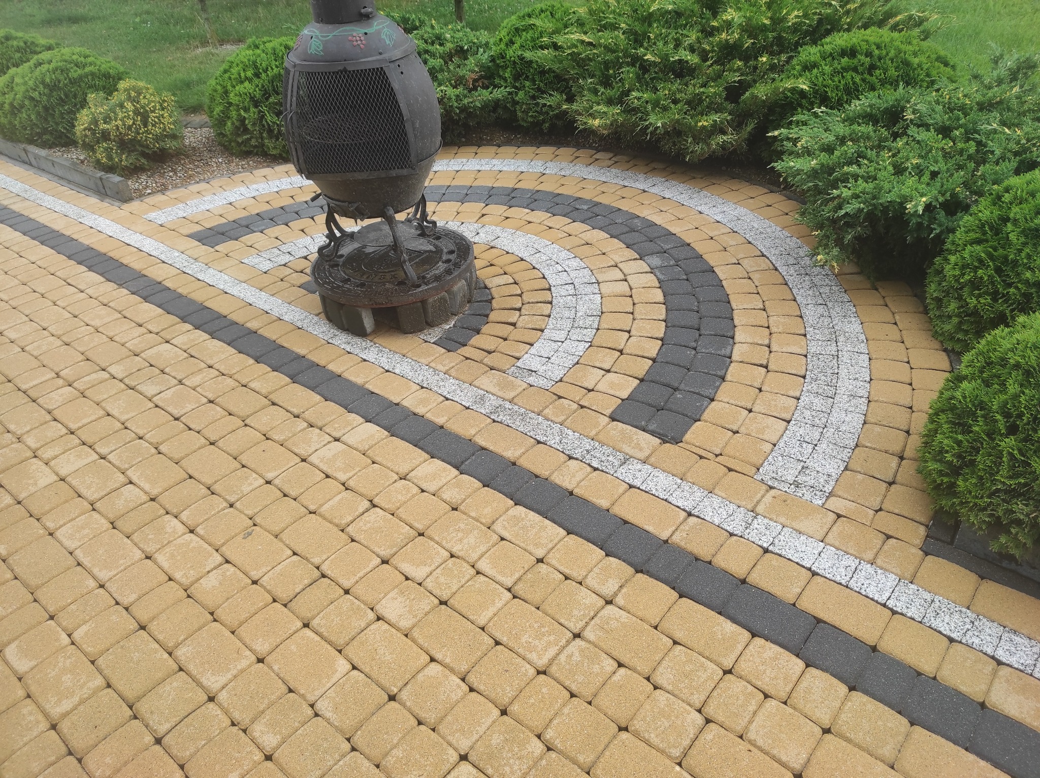 Ułożona kostka brukowa w odcieniach beżu, szarości i bieli tworząca wzór z okręgami, obok dekoracyjny metalowy kosz na ogień i zielone krzewy.