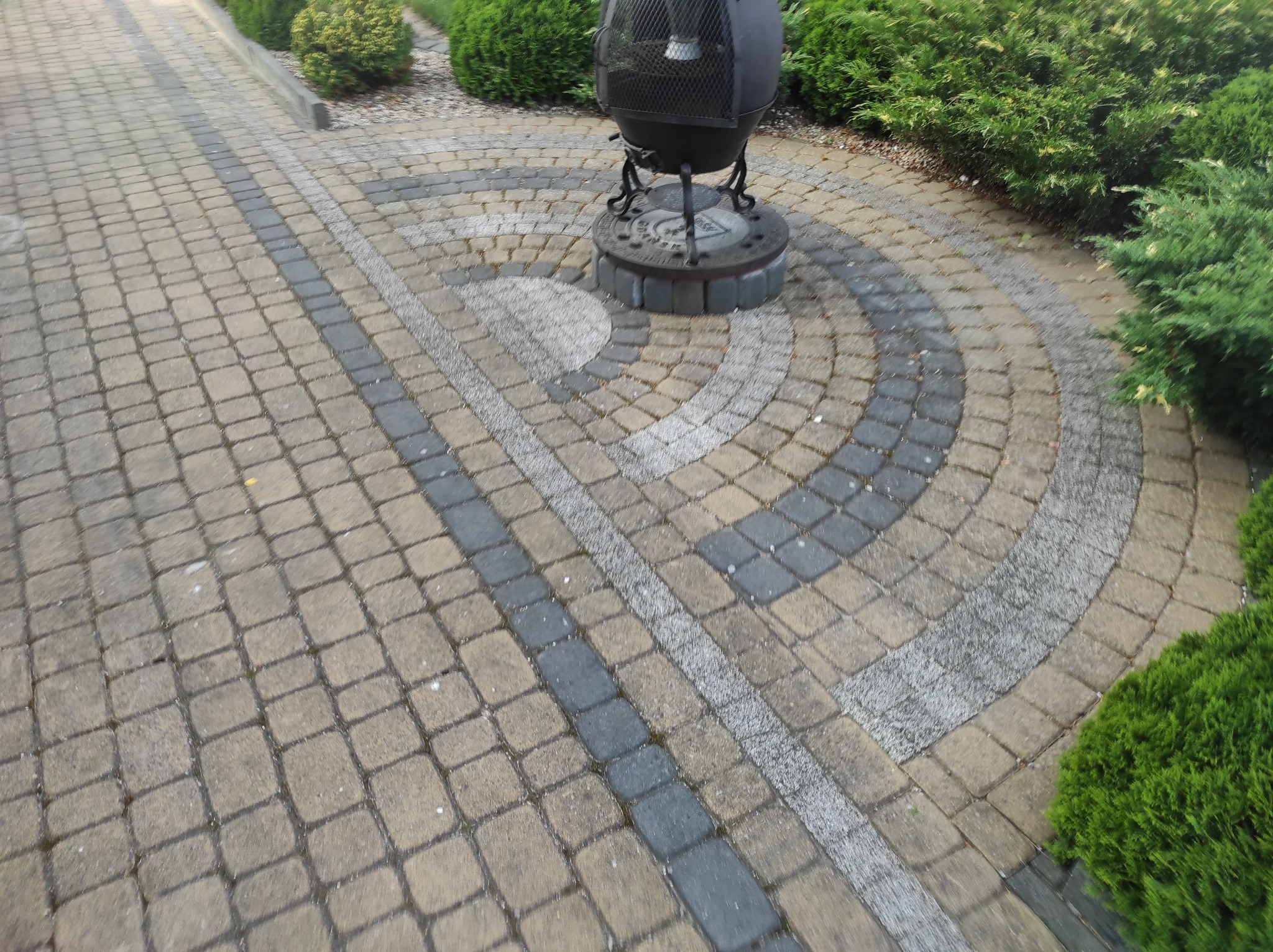 Ułożona z kostki brukowej ścieżka ogrodowa z elementami dekoracyjnymi w postaci okręgów z jasnej kostki i rzędu ciemnej kostki, zakończona okrągłym paleniskiem ogrodowym z metalową osłoną.