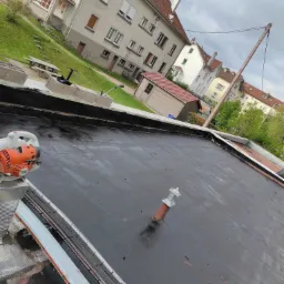 Czarny, płaski dach pokryty papą, z widocznym kominkiem wentylacyjnym i dmuchawą Stihl na krawędzi, na tle zabudowy mieszkalnej w pochmurny dzień.