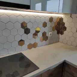 Kuchnia z heksagonalnymi płytkami w odcieniach szarości i złota, podświetlona listwą LED, z widoczną płytą indukcyjną i fragmentem blatu.